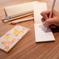 Person writing on a notepad with a pencil, next to a floral notebook and stationery items on a wooden surface.